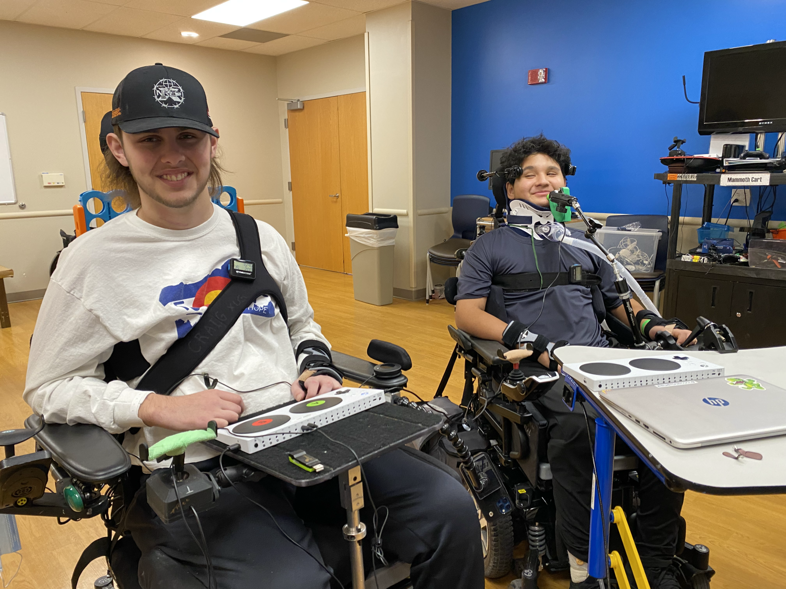 Two wheelchair users smile at the camera in front of their accessible gaming setups 